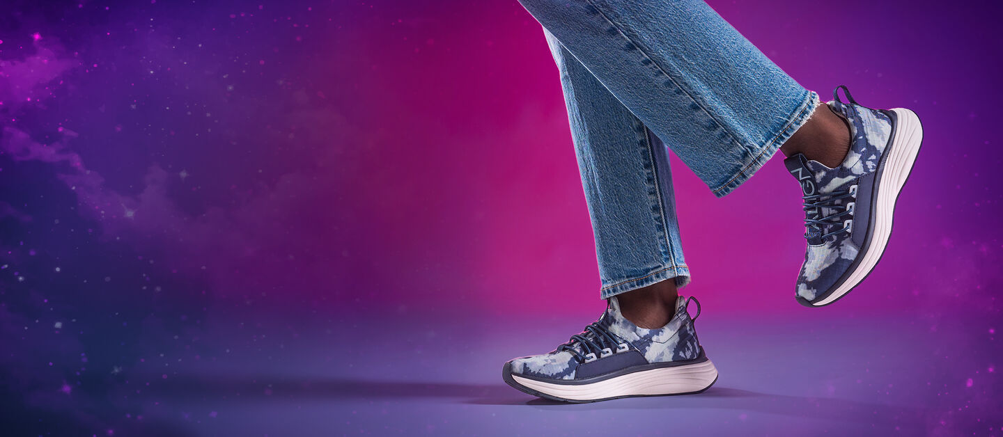 Women’s Theora Sneakers in Blue Tie Dye on foot on a pink and purple background with the back foot in the air